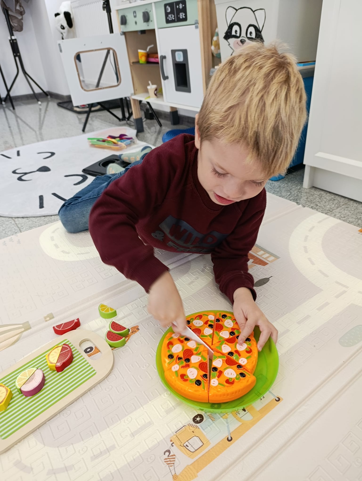 Chłopiec kroi zabawkę - pizzę, leżąc na macie edukacyjnej w pokoju zabaw. W tle widoczna dziecięca kuchnia i pluszowy miś panda.