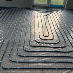 Niebieskie rury ogrzewania podłogowego ułożone na folii z czerwoną siatką, widok z góry, częściowo oświetlone przez okno.