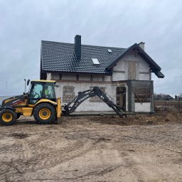 MPRO - Żółto-czarna koparko-ładowarka Volvo BL71 wykonuje prace ziemne przy budowie domu jednorodzinnego z szarą dachówką, w tle widoczny wiatrak.