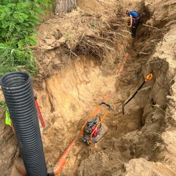 MPRO - Głęboki wykop w ziemi z widocznymi pomarańczowymi rurami kanalizacyjnymi, czarną rurą karbowaną i zagęszczarką spalinową. W tle pracownik w wykopie i ciężki sprzęt budowlany.