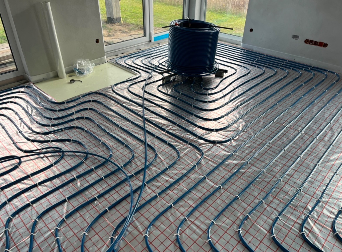 Instalacja ogrzewania podłogowego w trakcie montażu: widoczne niebieskie rurki ułożone na folii z siatką, zwoje rur na podłodze, surowy stan wykończenia wnętrza z oknami i fragmentami ścian.