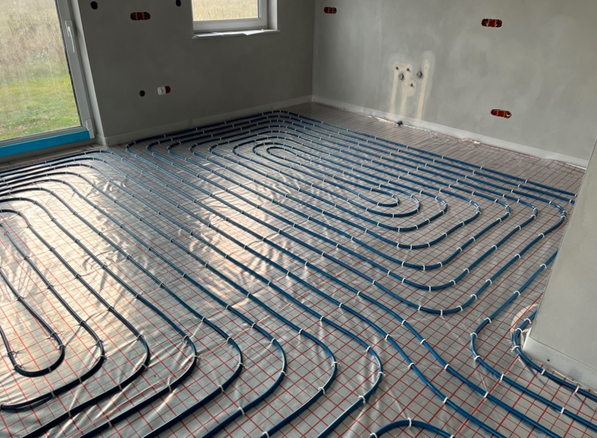Niebieskie rurki ogrzewania podłogowego ułożone w pętle na folii z czerwoną kratką w surowym wnętrzu, widoczne otwory na gniazdka elektryczne w ścianie.