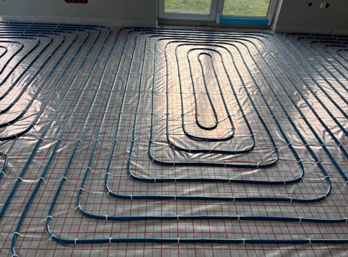 Niebieskie rury ogrzewania podłogowego ułożone na folii z czerwoną siatką, widok z góry, częściowo oświetlone przez okno.