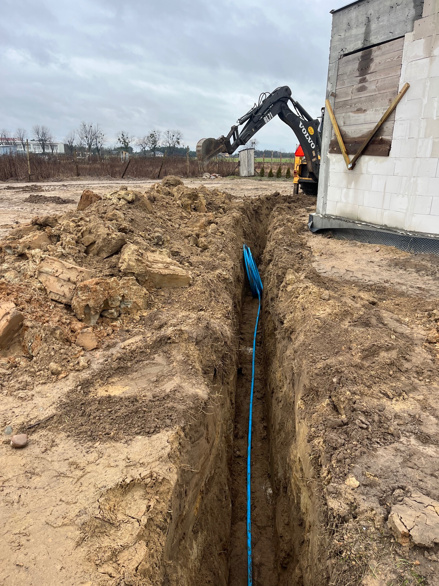 Wykopany rów z niebieską rurą w ziemi, w tle koparka Volvo i budynek w trakcie budowy.