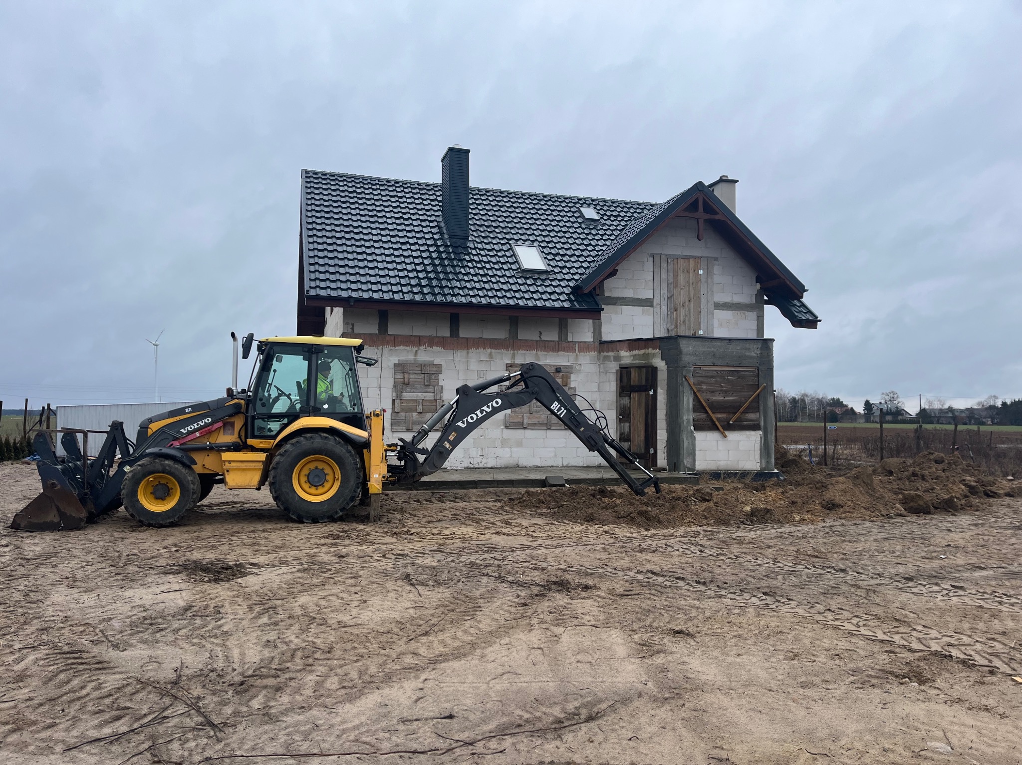Żółto-czarna koparko-ładowarka Volvo BL71 wykonuje prace ziemne przy budowie domu jednorodzinnego z szarą dachówką, w tle widoczny wiatrak.
