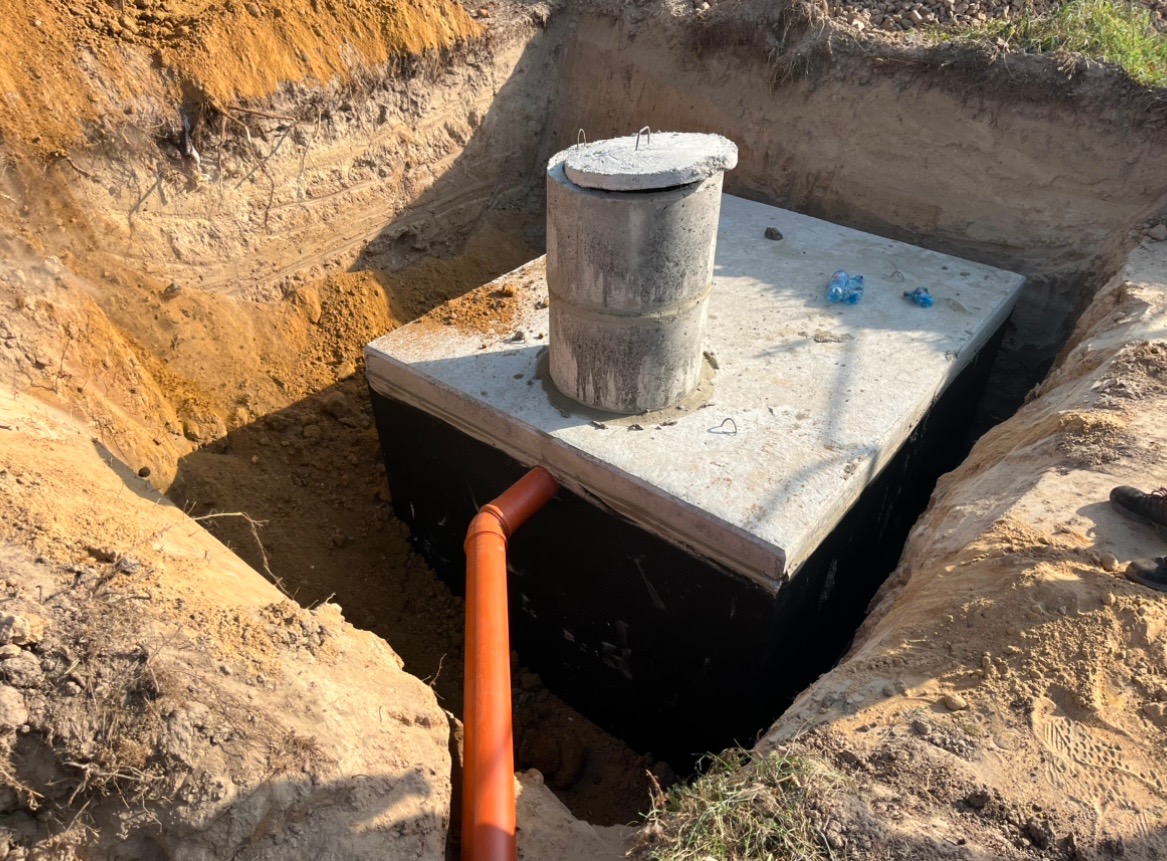 Betonowy zbiornik przydomowej oczyszczalni ścieków w wykopie, zabezpieczony czarną folią, z podłączoną pomarańczową rurą kanalizacyjną.