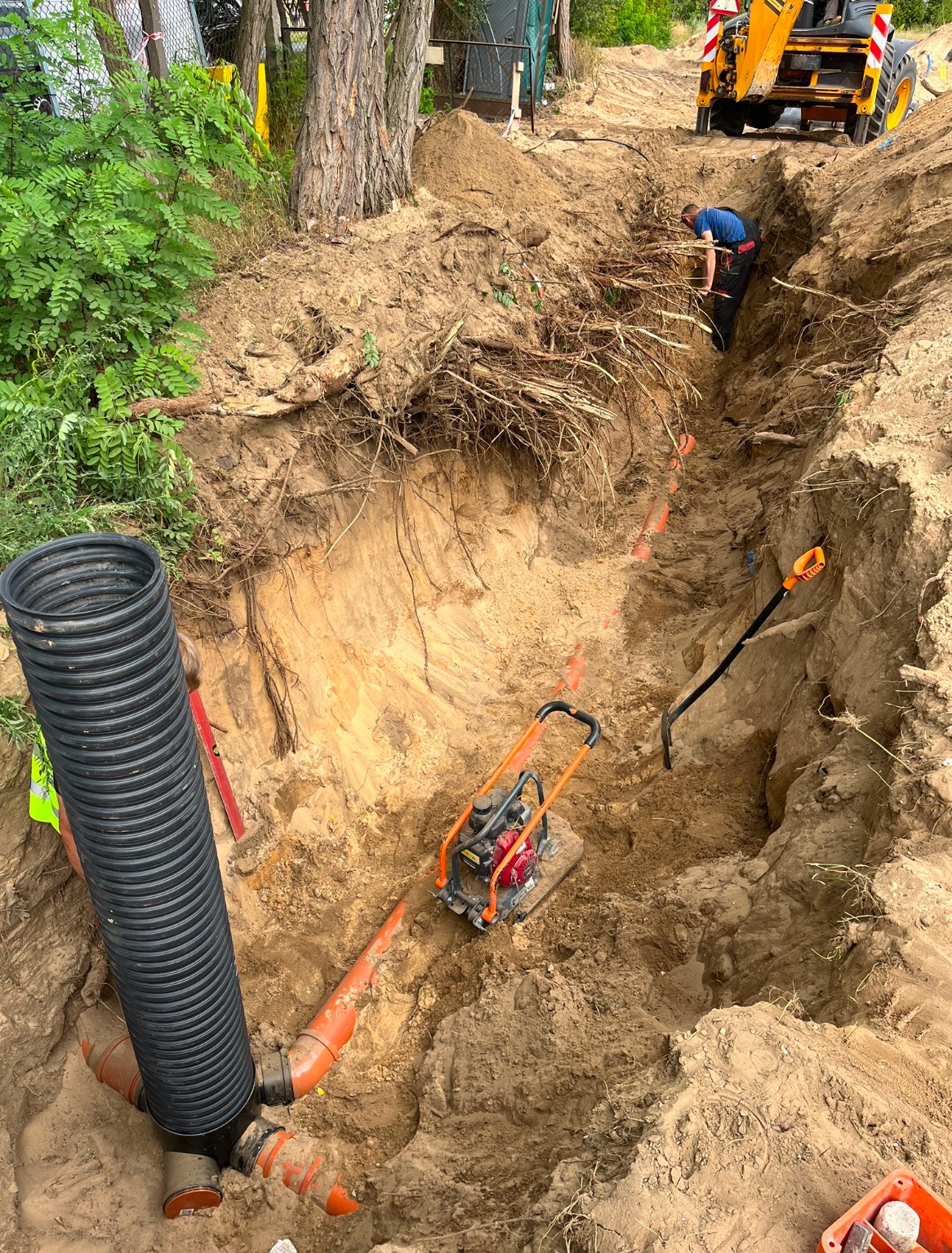 Głęboki wykop w ziemi z widocznymi pomarańczowymi rurami kanalizacyjnymi, czarną rurą karbowaną i zagęszczarką spalinową. W tle pracownik w wykopie i ciężki sprzęt budowlany.