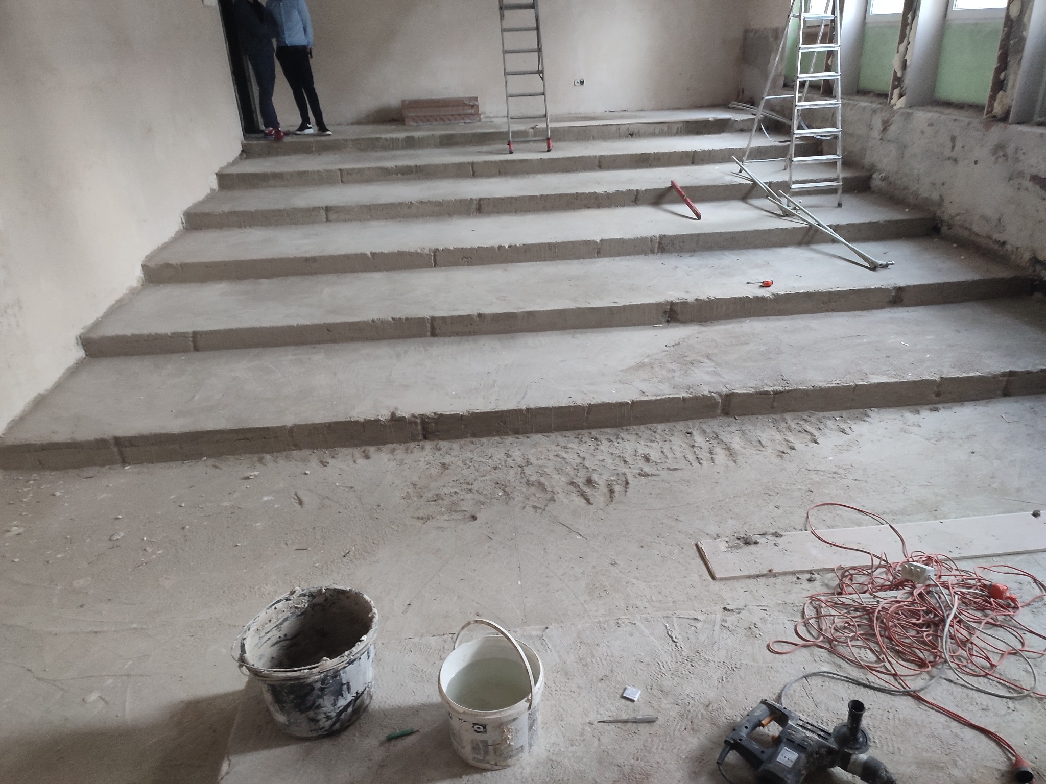 Wnętrze w trakcie prac budowlanych: surowe, betonowe schody, dwa wiadra, narzędzia, kable i drabiny. Widoczne ślady po pracach wykończeniowych.