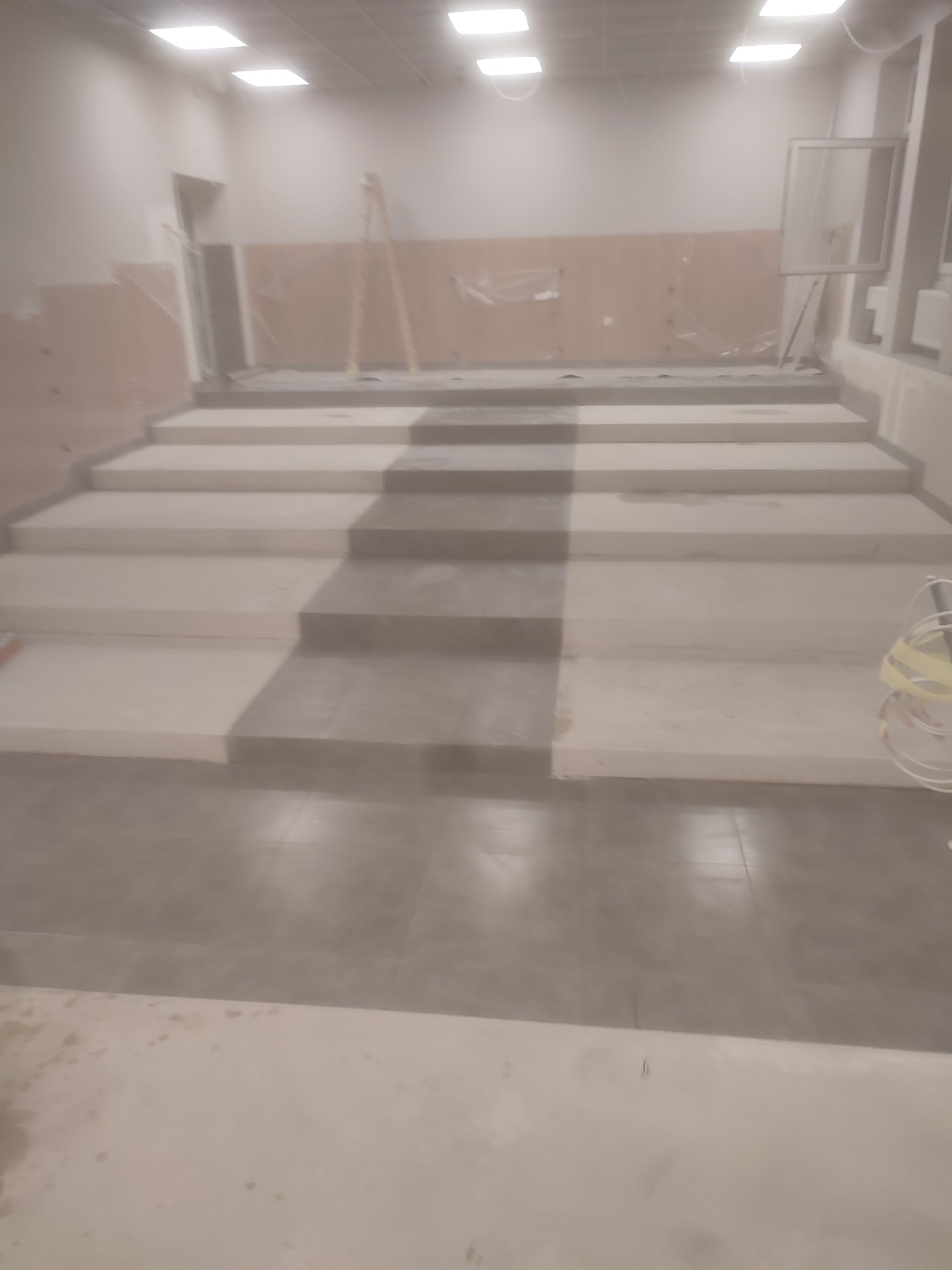 Wnętrze w trakcie prac wykończeniowych: ułożone płytki podłogowe w szarym odcieniu, częściowo wykończone schody z widocznymi śladami po szpachlowaniu, przygotowane ściany i sufity, widoczne...