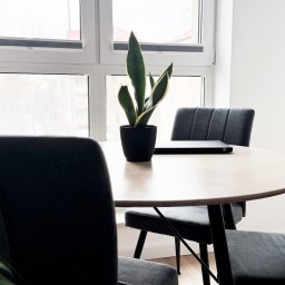 Złota 2 Sp. z o.o. - Jasne wnętrze biurowe z okrągłym stołem, szarymi krzesłami i rośliną w doniczce, laptop na stole, widok na okno.