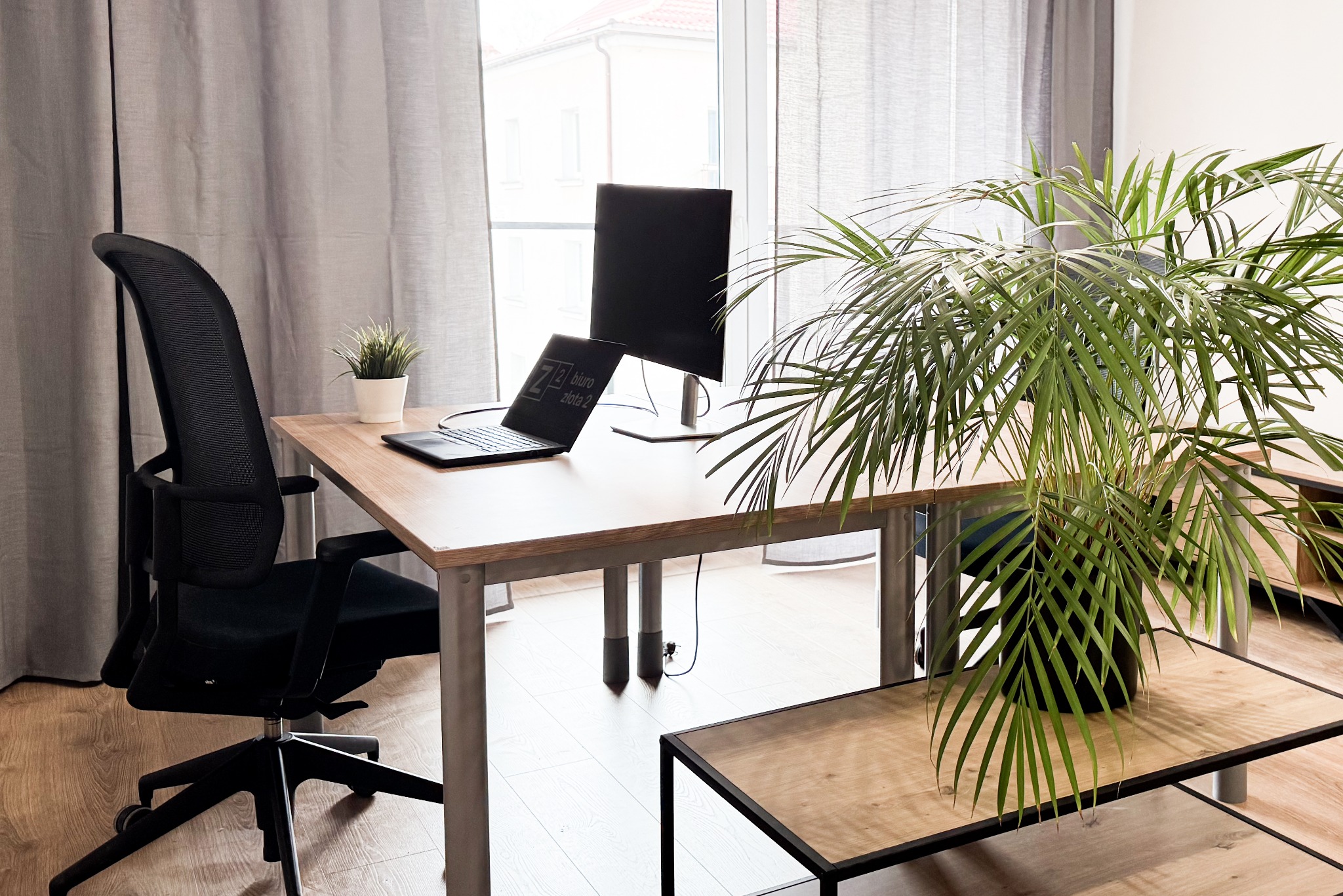 Jasne biuro z drewnianym biurkiem, czarnym krzesłem biurowym, laptopem z widocznym logo 'Z2 Biuro Złota 2' na ekranie, monitorem, roślinami doniczkowymi i szarymi zasłonami w tle.