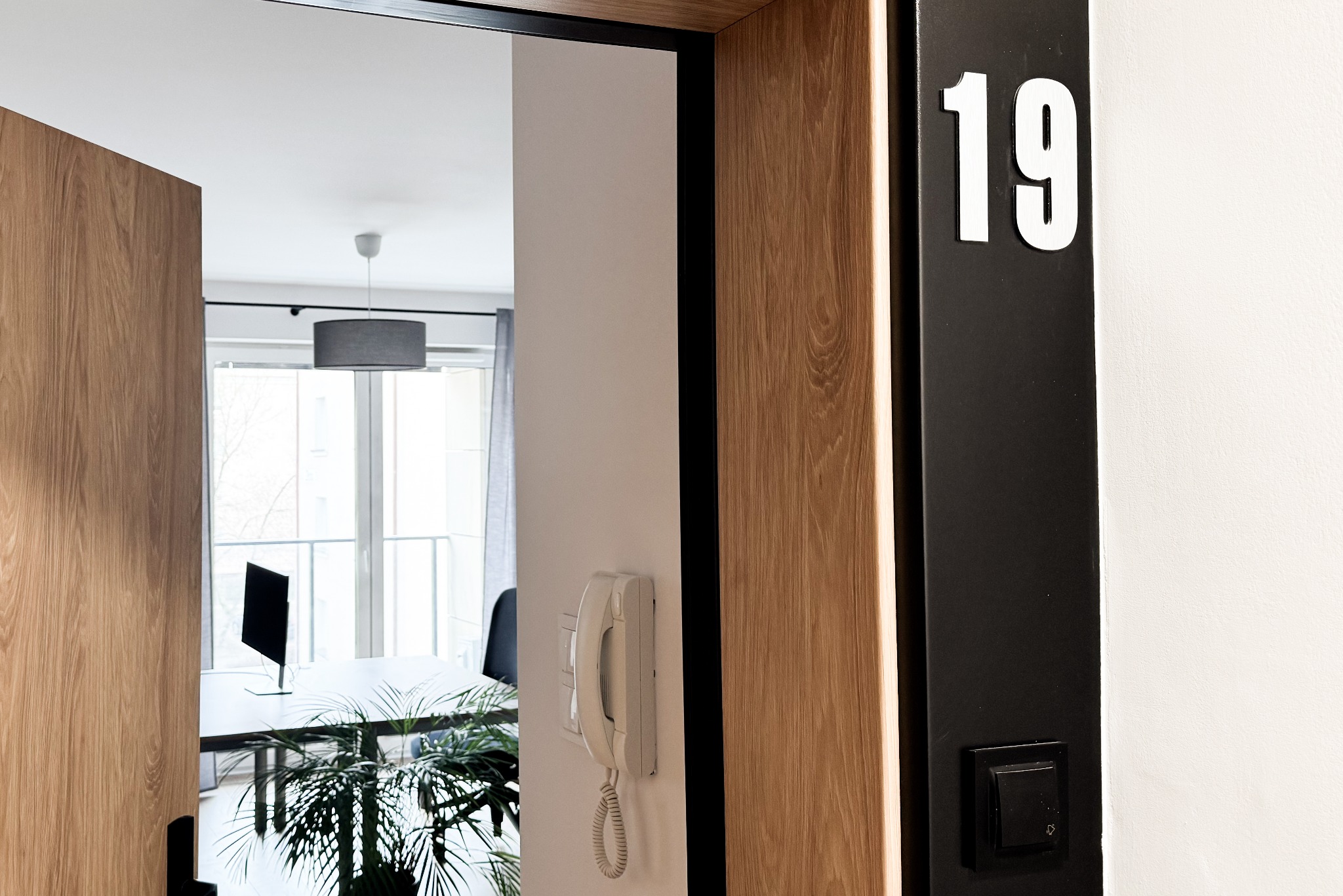 Wejście do biura numer 19 z widocznym biurkiem, komputerem i rośliną doniczkową w tle, telefon na ścianie.