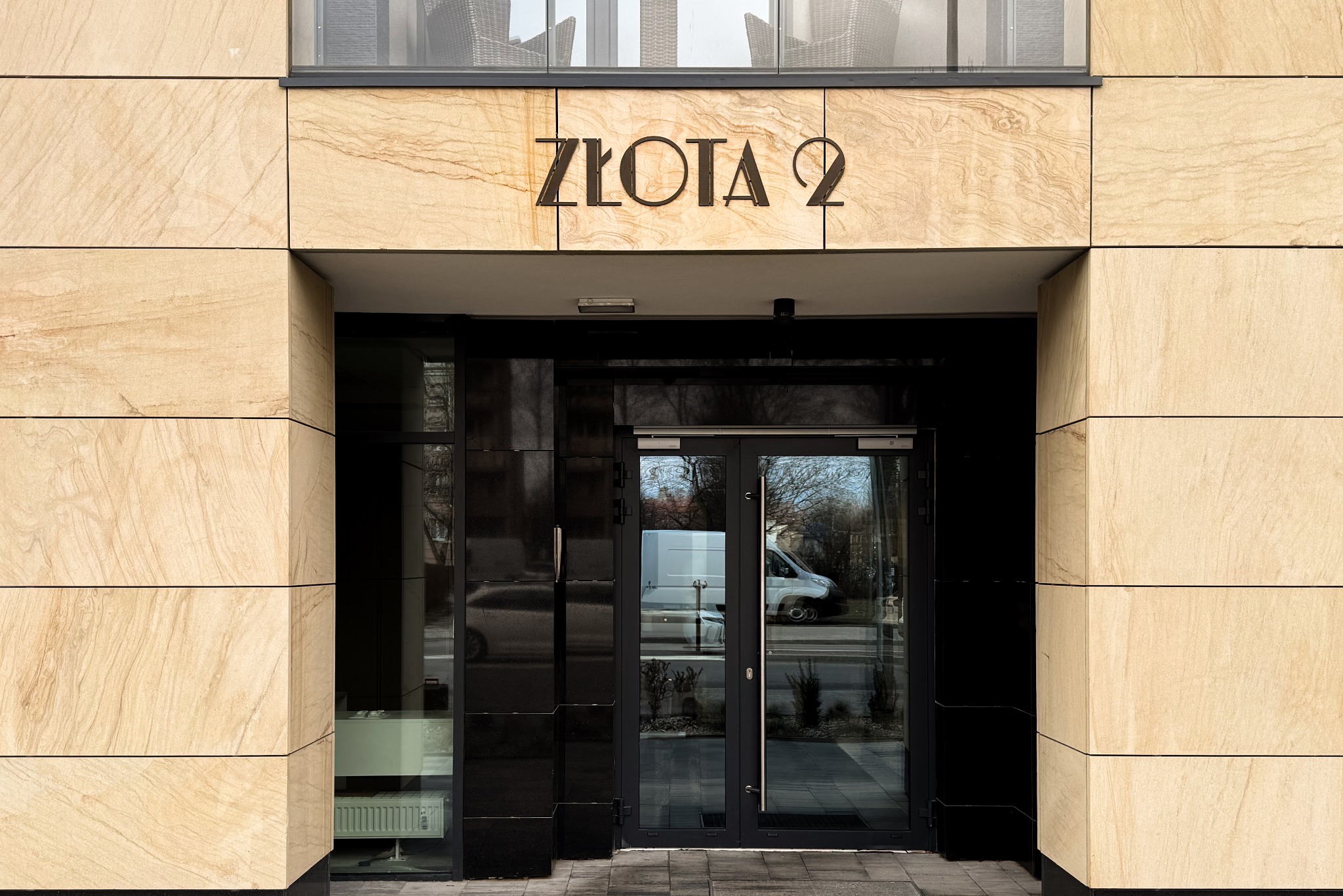 Wejście do budynku z adresem 'Złota 2' nad drzwiami, widoczne odbicie białego vana w szklanych drzwiach.