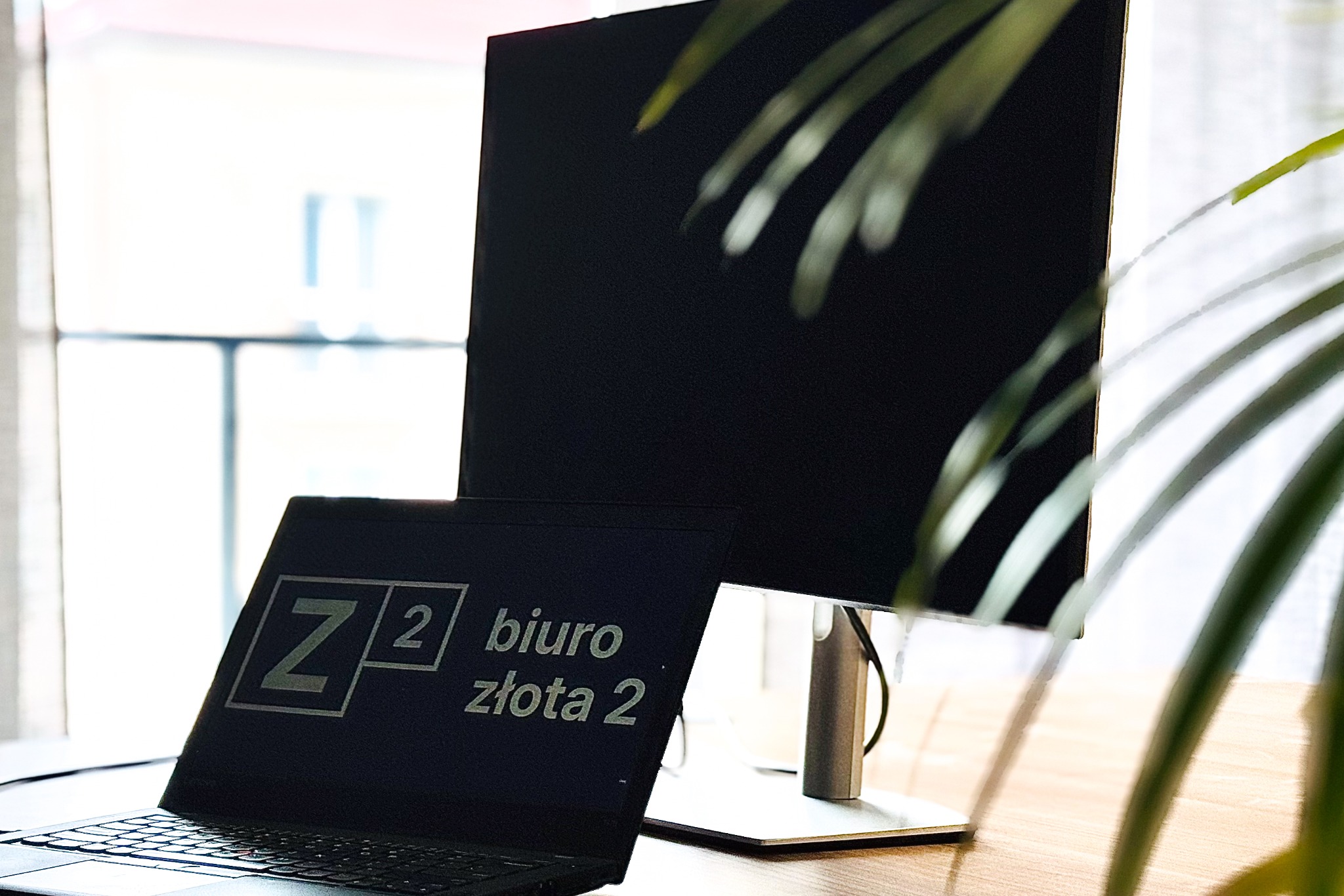 Laptop z logo 'Z2 biuro złota 2' na tle monitora i rośliny doniczkowej w Białymstoku, widok z bliska.