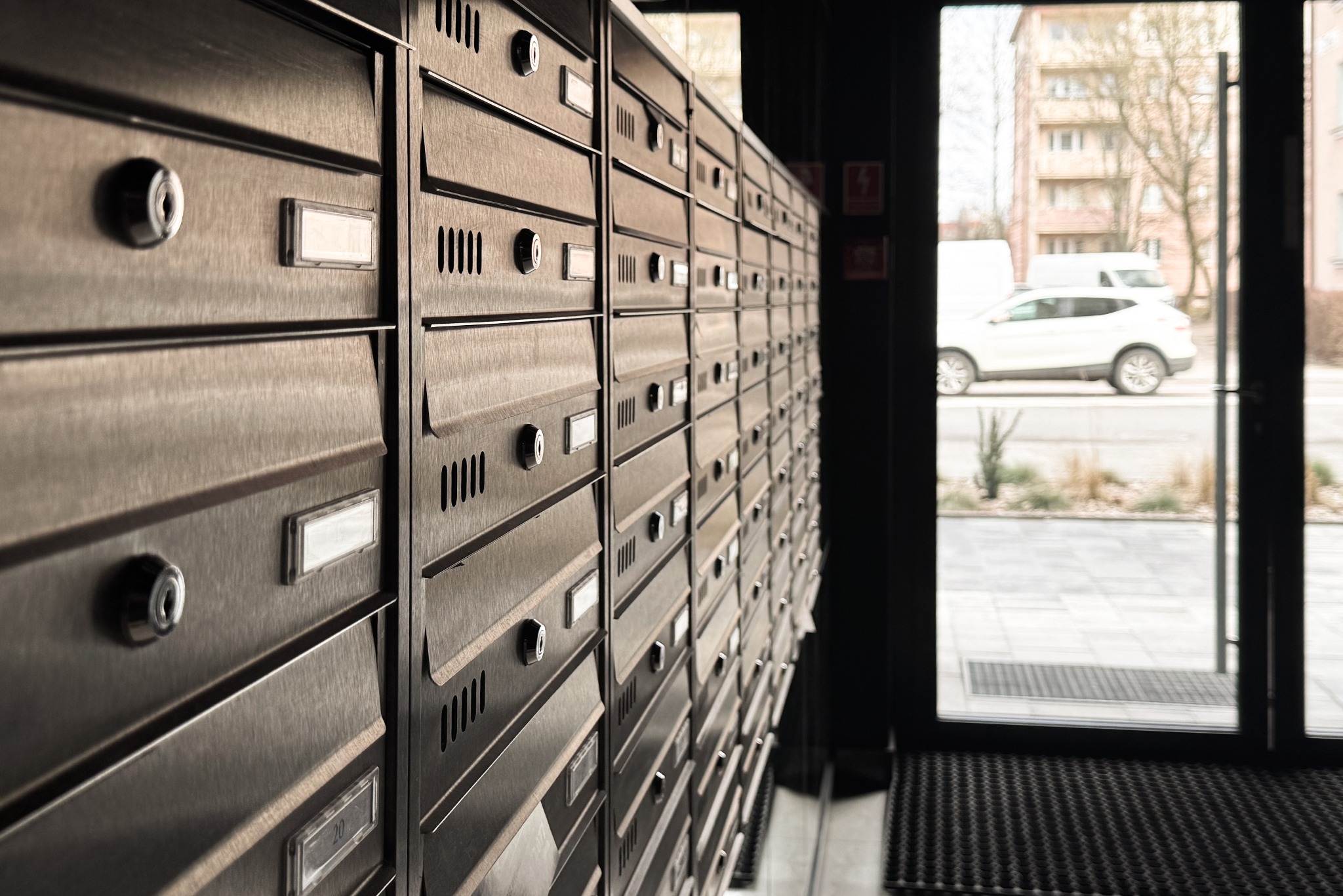 Rząd metalowych skrytek pocztowych wewnątrz budynku, widok przez szklane drzwi na ulicę z zaparkowanym białym samochodem.