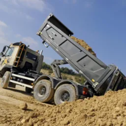 Ciężarówka wywrotka z podniesioną skrzynią ładunkową, wysypująca piasek na ziemię. Widok z dołu na tle błękitnego nieba.