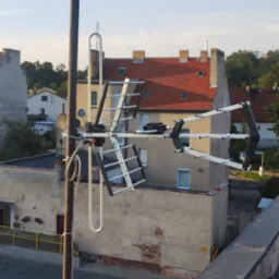 Antena DVB-T na dachu budynku. Widok na okoliczne budynki i niebo. Antena zamontowana na metalowym maszcie, białe kable przymocowane do masztu.