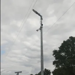 Słup z lampą LED i dwiema kamerami monitoringu na tle pochmurnego nieba i pola, widoczne ogrodzenie terenu.