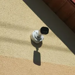 Kamera monitoringu zamontowana na elewacji budynku, z widocznym fragmentem dachu i niebem. Całość w słonecznym oświetleniu.