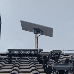 Strefa360-systemy zabezpieczeń - Kamera monitoringu zamontowana na dachu z czarnej dachówki, widok z dołu. Metalowa konstrukcja mocująca. Kabel zasilający widoczny. Szare niebo w tle.