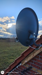 Antena satelitarna zamontowana na dachu z czerwonej cegły, widok z dołu, promienie słoneczne przebijają się przez chmury w tle.