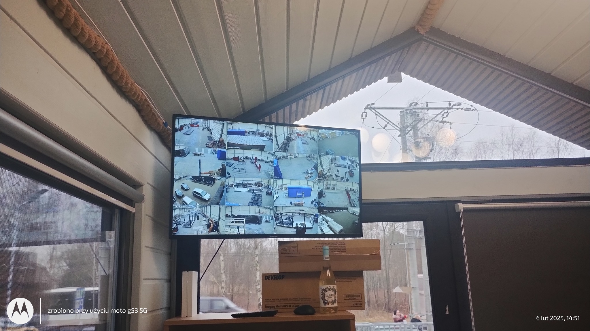 Ekran monitoringu CCTV z wieloma kamerami w pomieszczeniu, widok z wnętrza na zewnątrz przez okno, na pierwszym planie butelka wina i kartony.