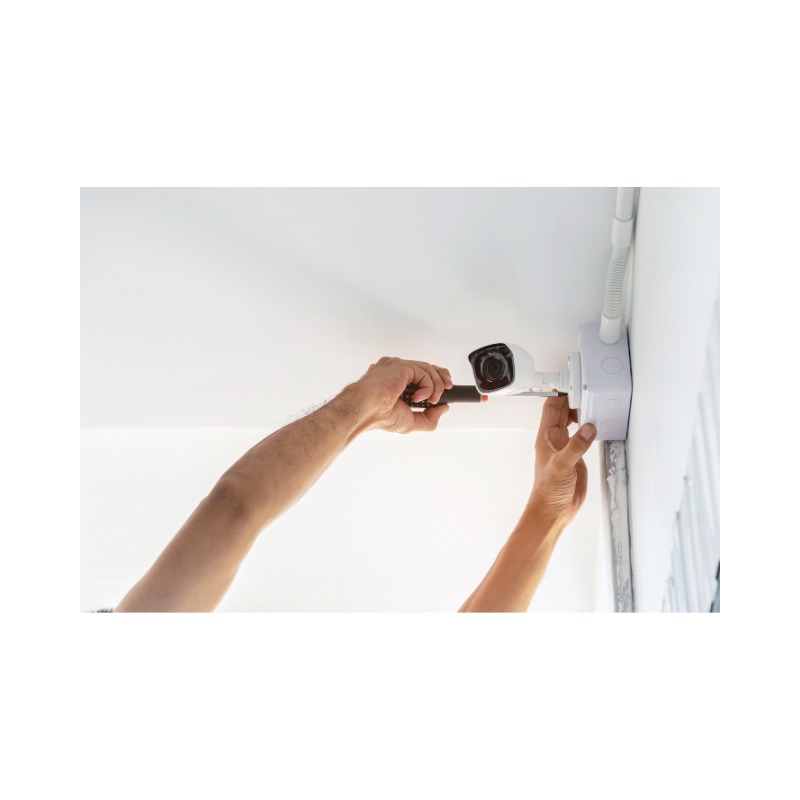 Instalacja kamery monitoringu: mężczyzna przykręca kamerę do puszki instalacyjnej na białym suficie, widoczne narzędzia i rury instalacyjne.