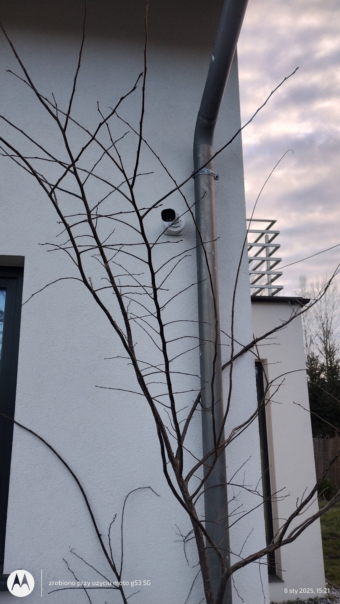Kamera monitoringu zewnętrznego zamontowana na elewacji budynku, obok rury spustowej, częściowo zasłonięta przez gałęzie nagiego drzewa.