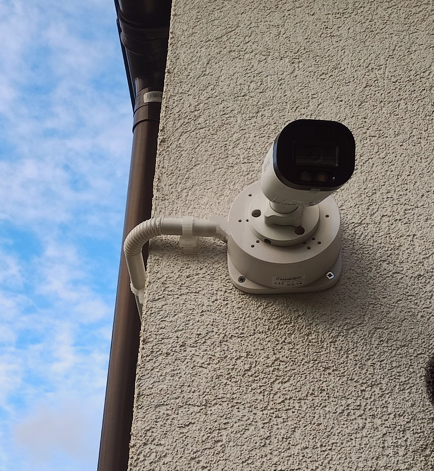 Zewnętrzna kamera monitoringu, zamontowana na teksturowanej ścianie budynku obok brązowej rynny, widok na błękitne niebo z chmurami.