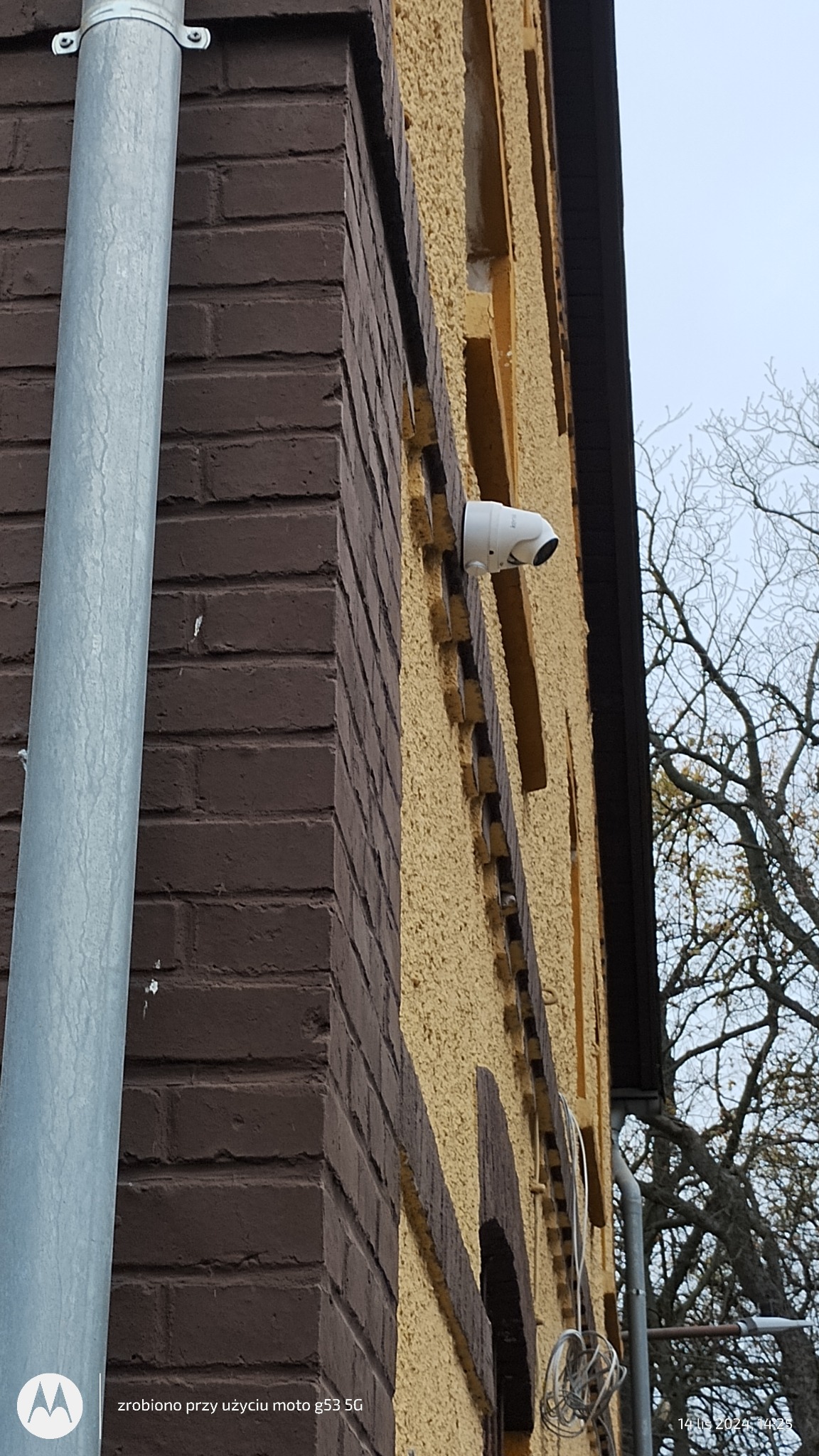 Zewnętrzna kamera monitoringu, biała, zamontowana na elewacji budynku z brązowej cegły i żółtym tynkiem strukturalnym, widok z dołu, obok rura spustowa.
