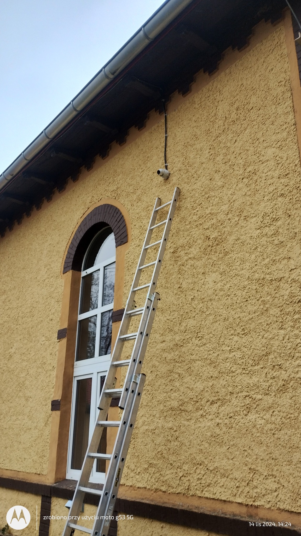 Kamera monitoringu zamontowana na elewacji budynku z drabiny przystawionej do ściany, widoczne okablowanie i charakterystyczna tekstura tynku.