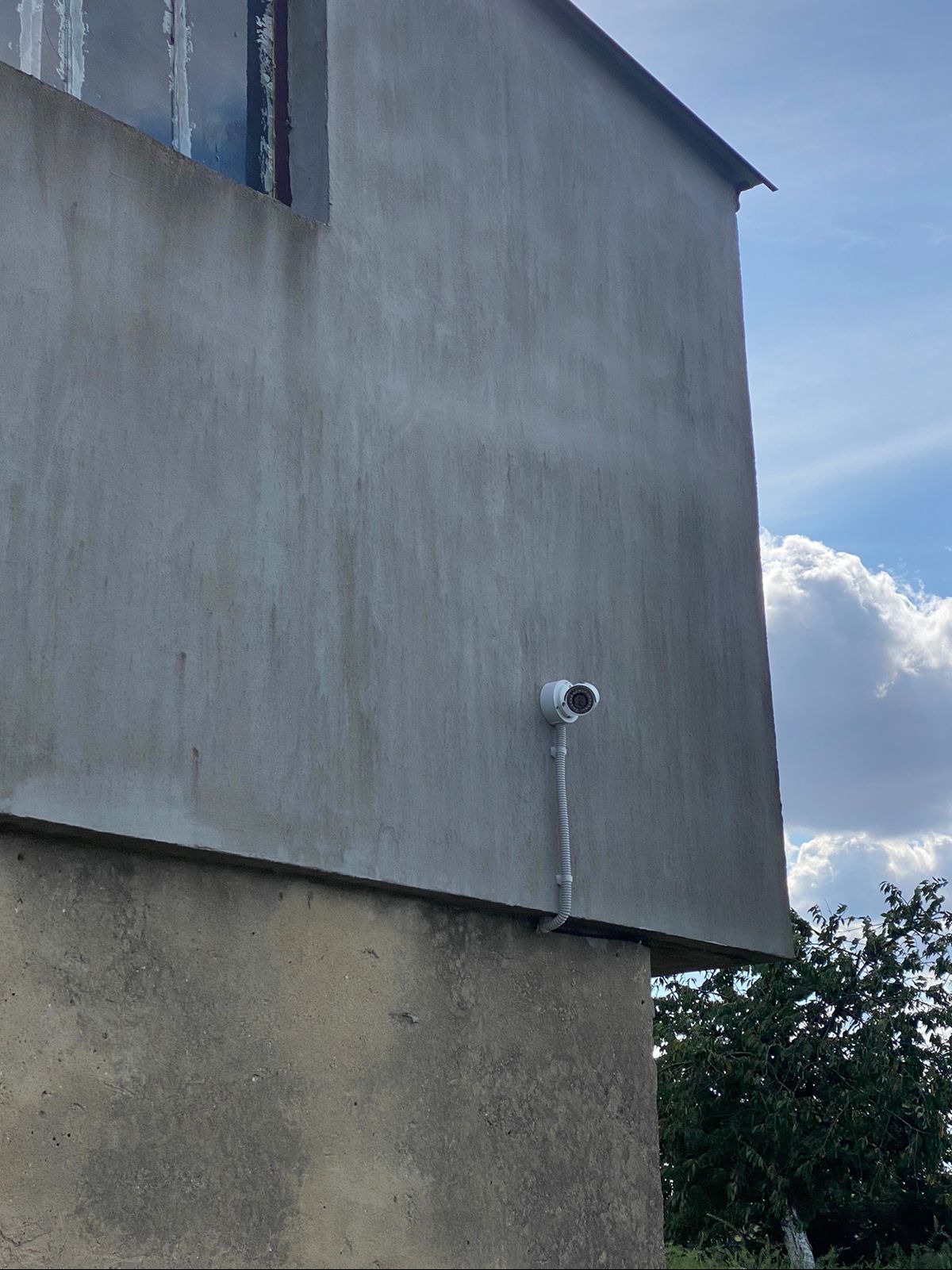 Biała kamera monitoringu zamontowana na szarej, betonowej ścianie budynku, widoczne okno i niebo z chmurami w tle.