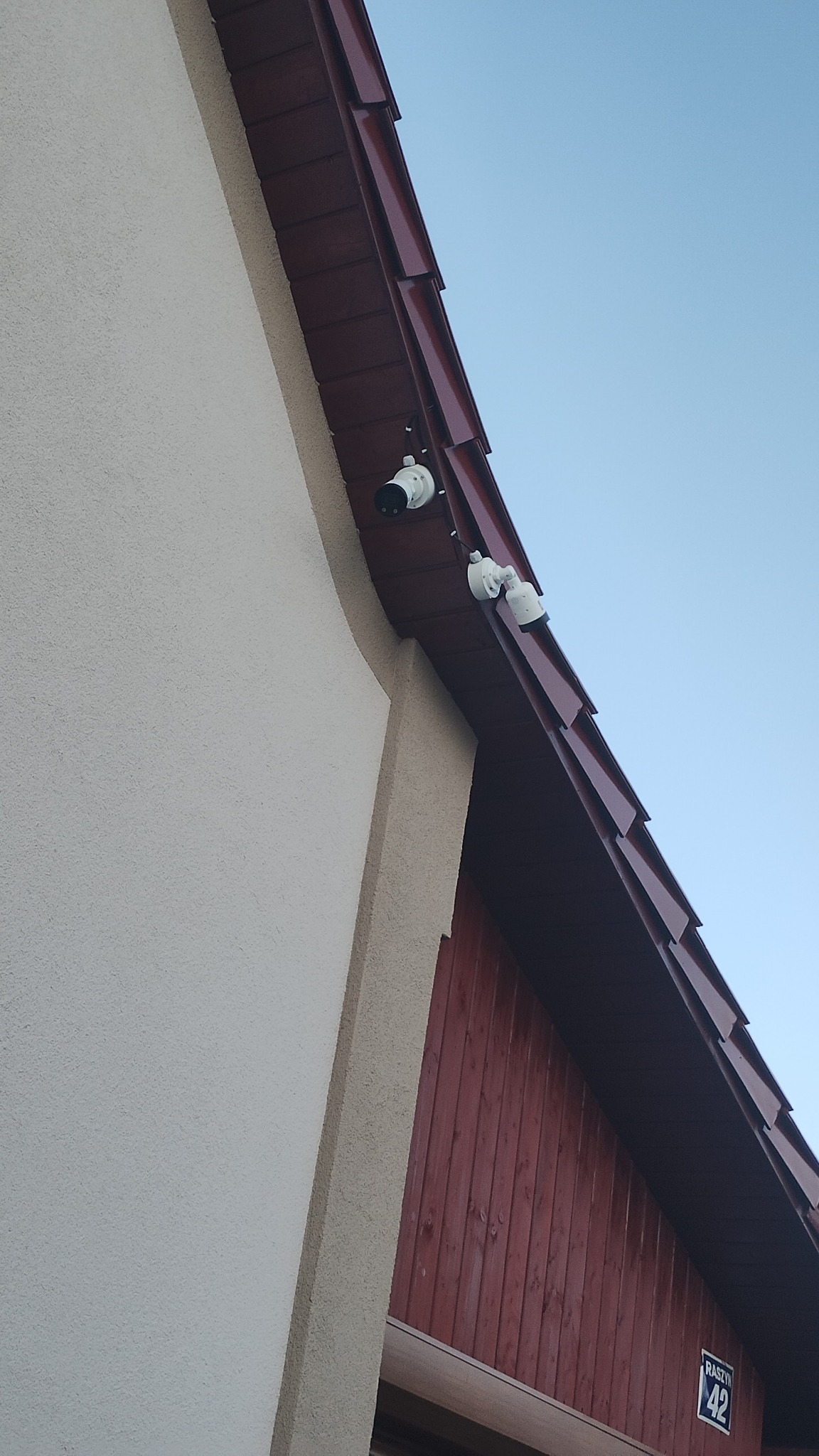 Dwie białe kamery monitoringu zamontowane pod okapem budynku z czerwonymi drewnianymi panelami, widoczne przewody, ujęcie z dołu na tle błękitnego nieba.