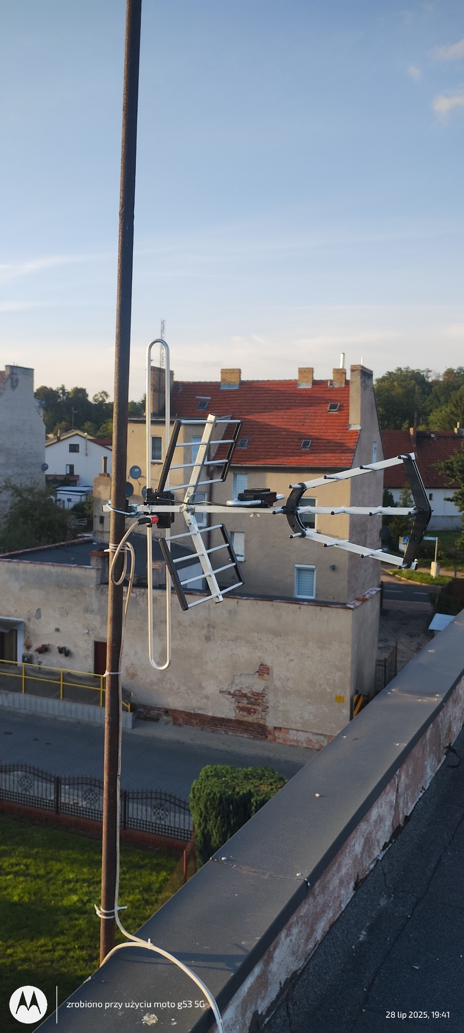 Antena DVB-T na dachu budynku. Widok na okoliczne budynki i niebo. Antena zamontowana na metalowym maszcie, białe kable przymocowane do masztu.