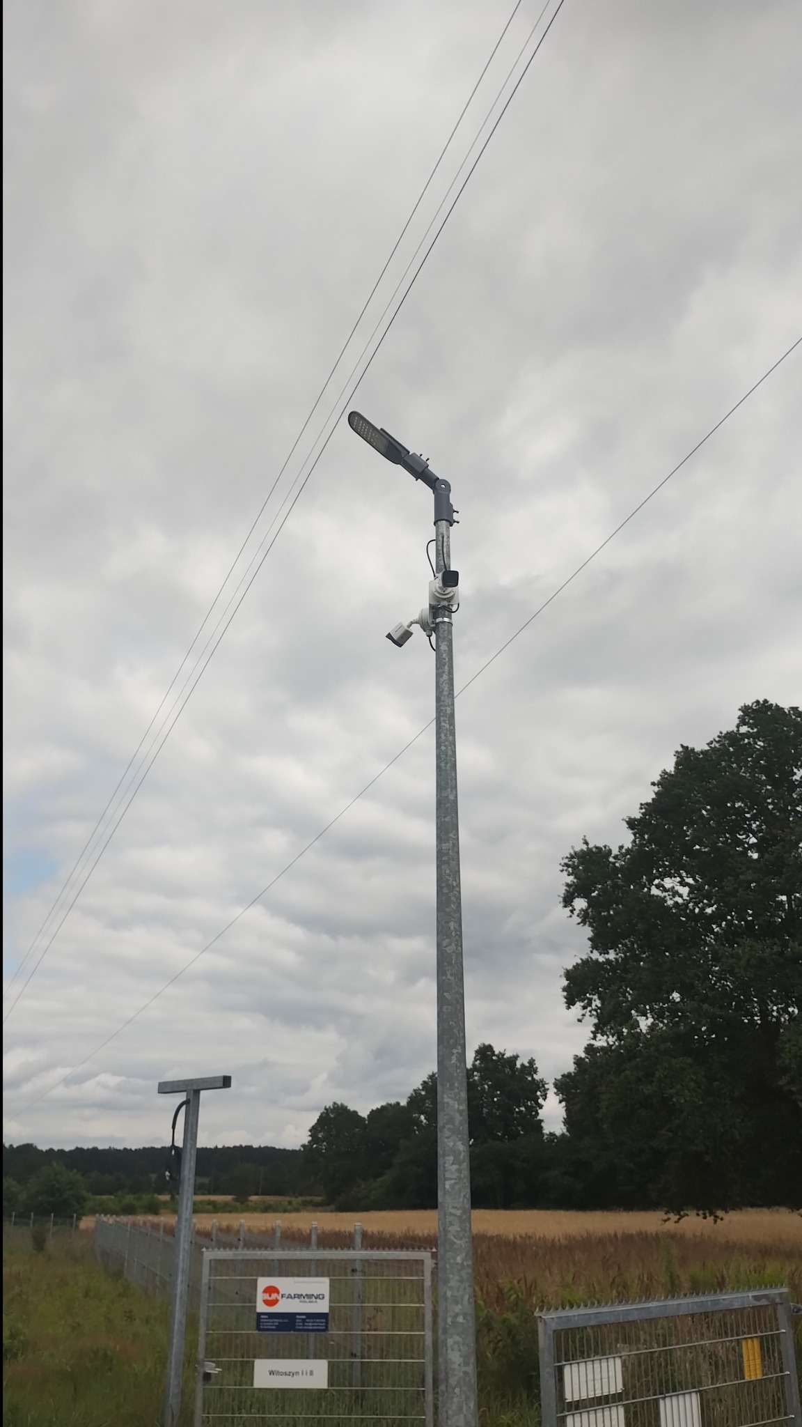 Słup z lampą LED i dwiema kamerami monitoringu na tle pochmurnego nieba i pola, widoczne ogrodzenie terenu.