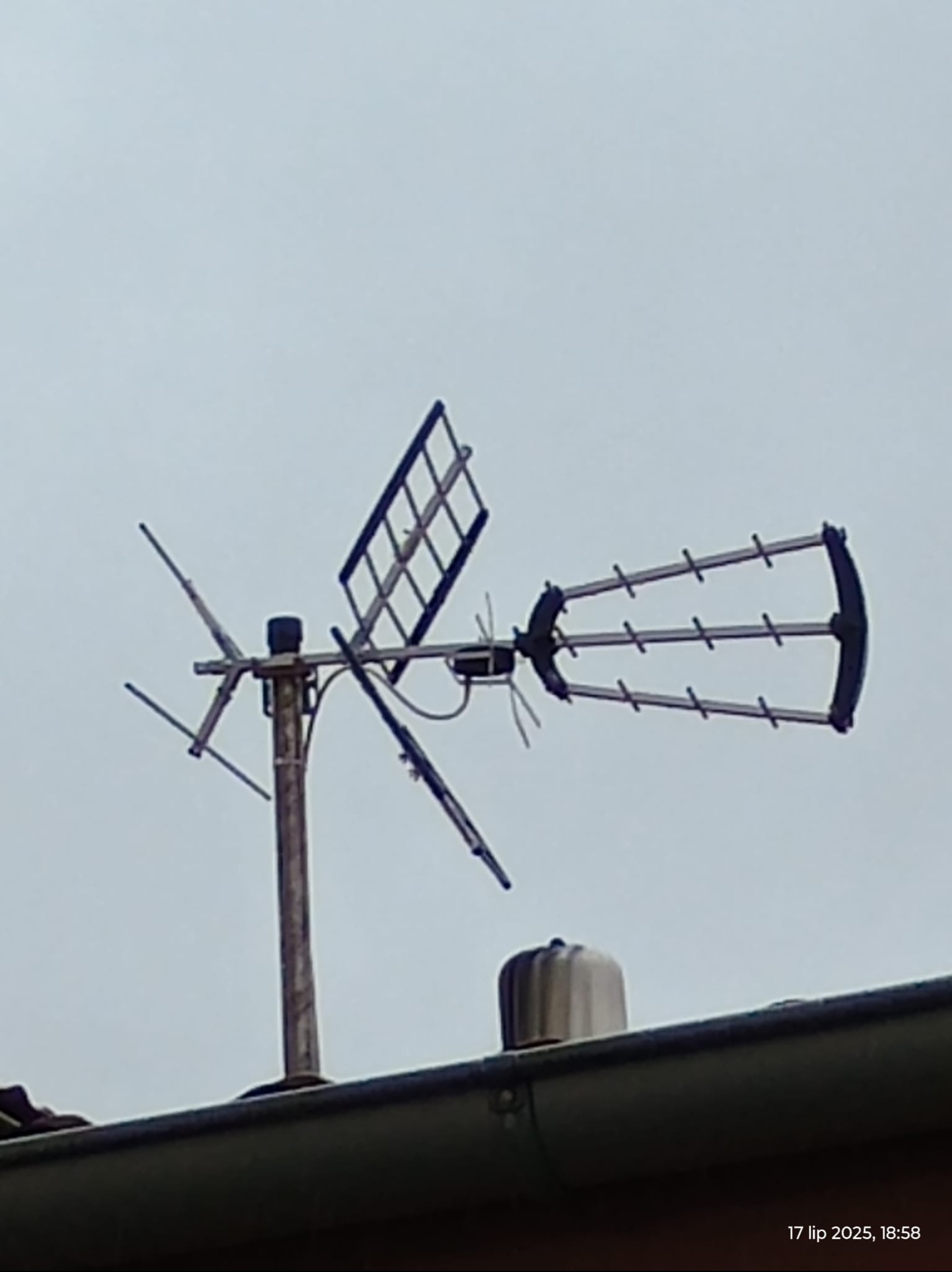 Antena telewizyjna typu Yagi-Uda zamontowana na dachu domu, widoczna z dołu na tle szarego nieba. Antena skierowana w lewo, z widocznymi elementami wzmacniającymi sygnał.