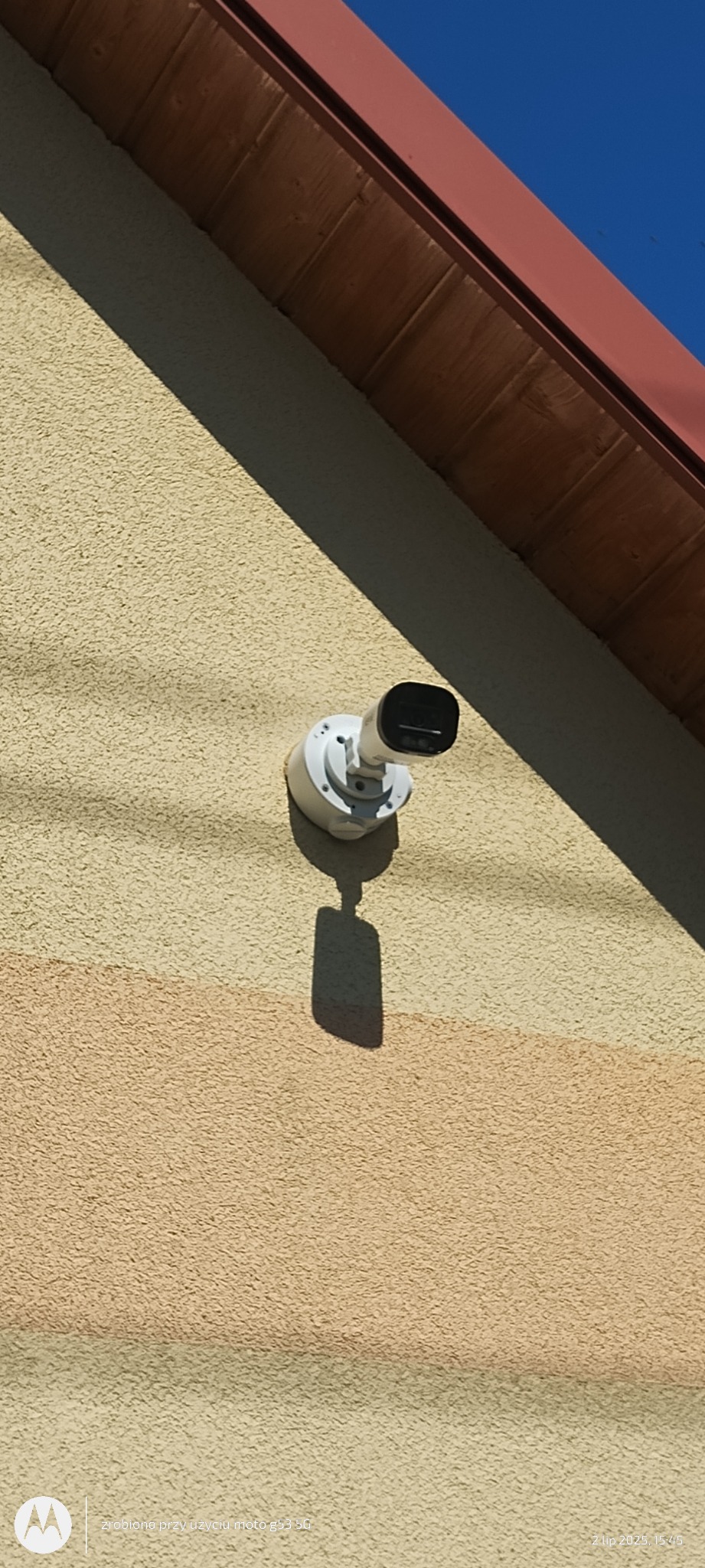 Kamera monitoringu zamontowana na elewacji budynku, z widocznym fragmentem dachu i niebem. Całość w słonecznym oświetleniu.