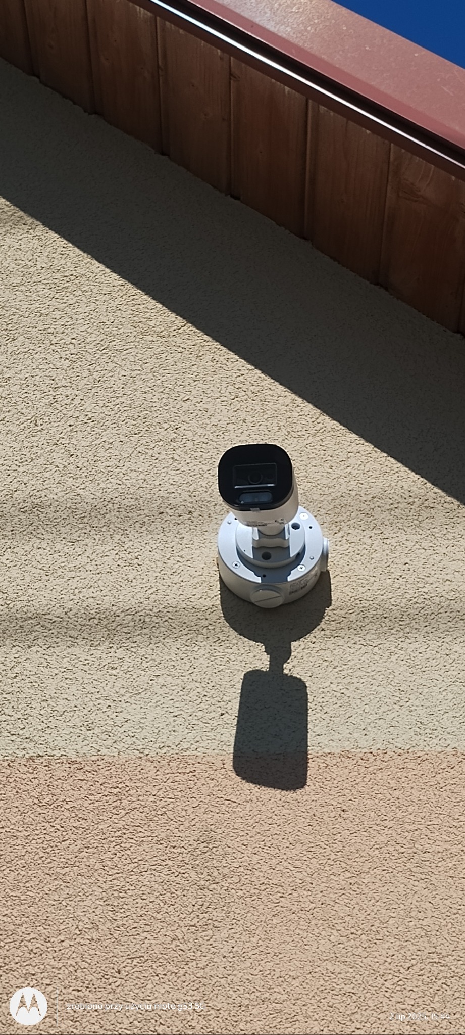 Kamera monitoringu IP zamontowana na elewacji budynku. Widoczny cień kamery na teksturowanej ścianie. Ujęcie z góry, podkreślające dyskretny montaż.