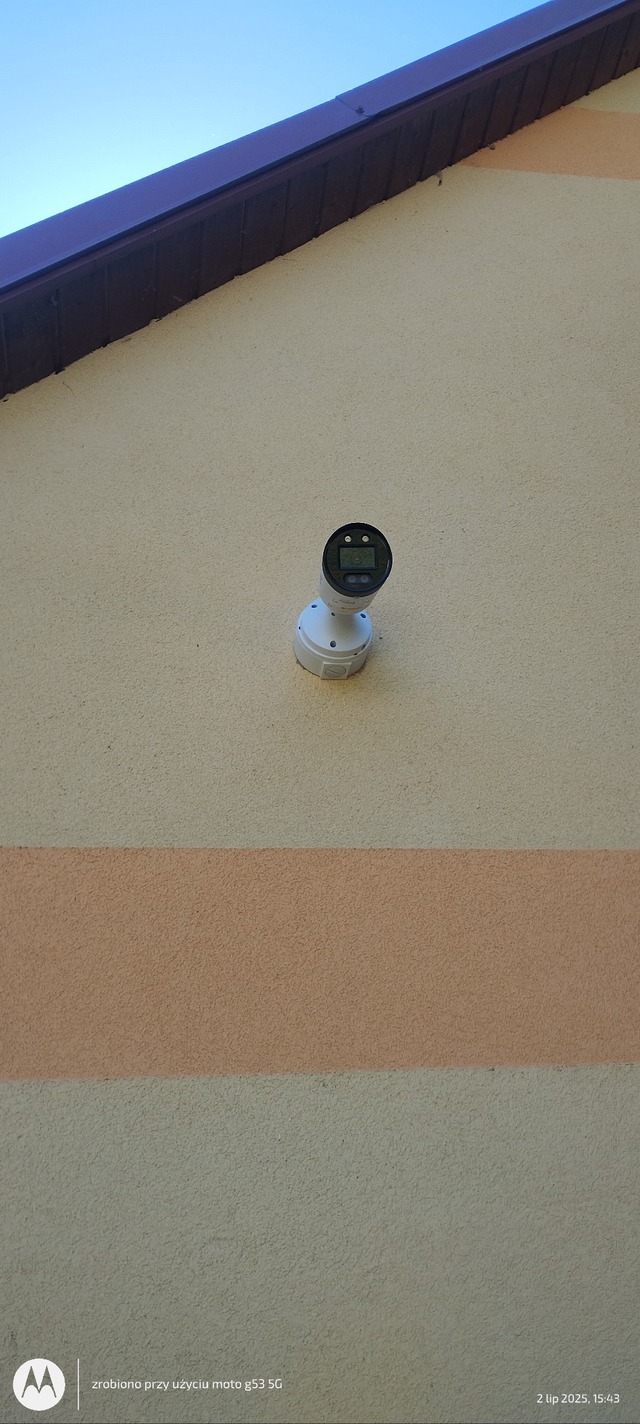 Kamera monitoringu, model kopułkowy, zamontowana na elewacji budynku z widocznym fragmentem dachu i niebem w tle. Jasna elewacja z poziomym pasem w odcieniu pomarańczowym.