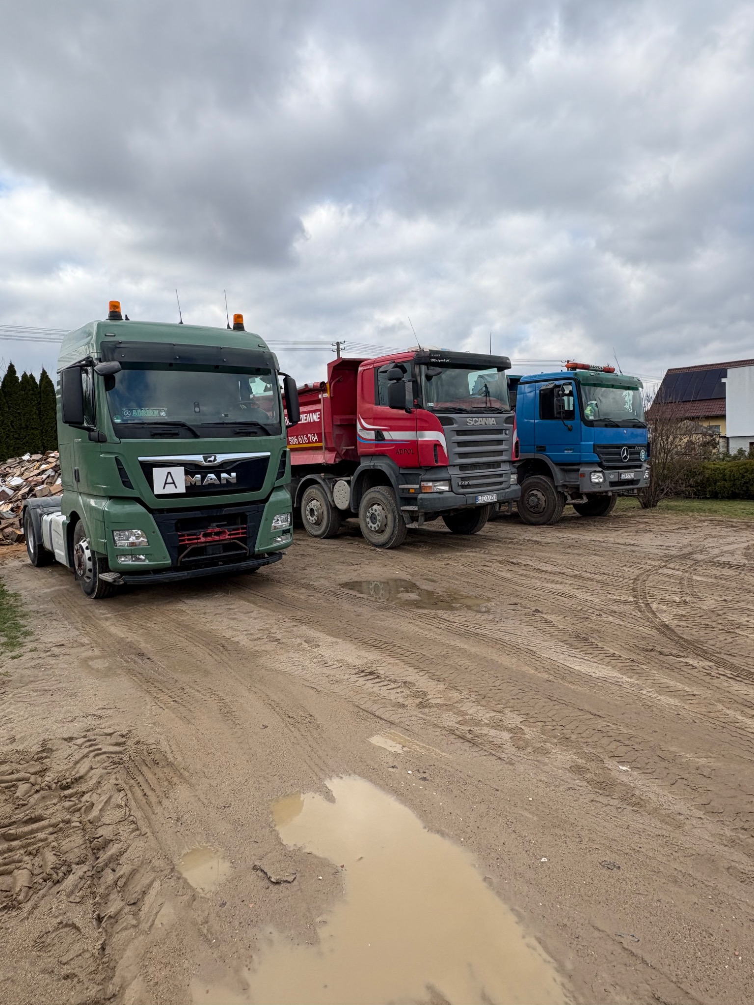 Trzy ciężarówki różnych marek (MAN, Scania, Mercedes) stojące na placu budowy po deszczu, widoczne ślady opon i kałuże na ziemi.