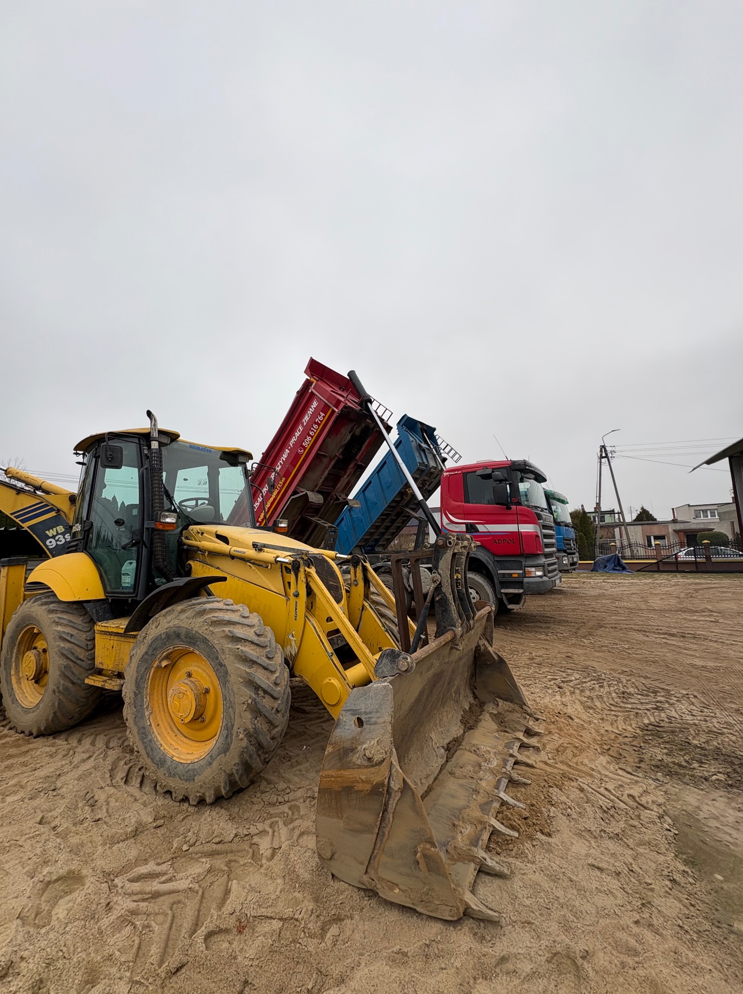 Żółta koparko-ładowarka Komatsu z łyżką na piasku, w tle wywrotki z podniesionymi skrzyniami ładunkowymi: czerwona, niebieska i zielona, szare niebo