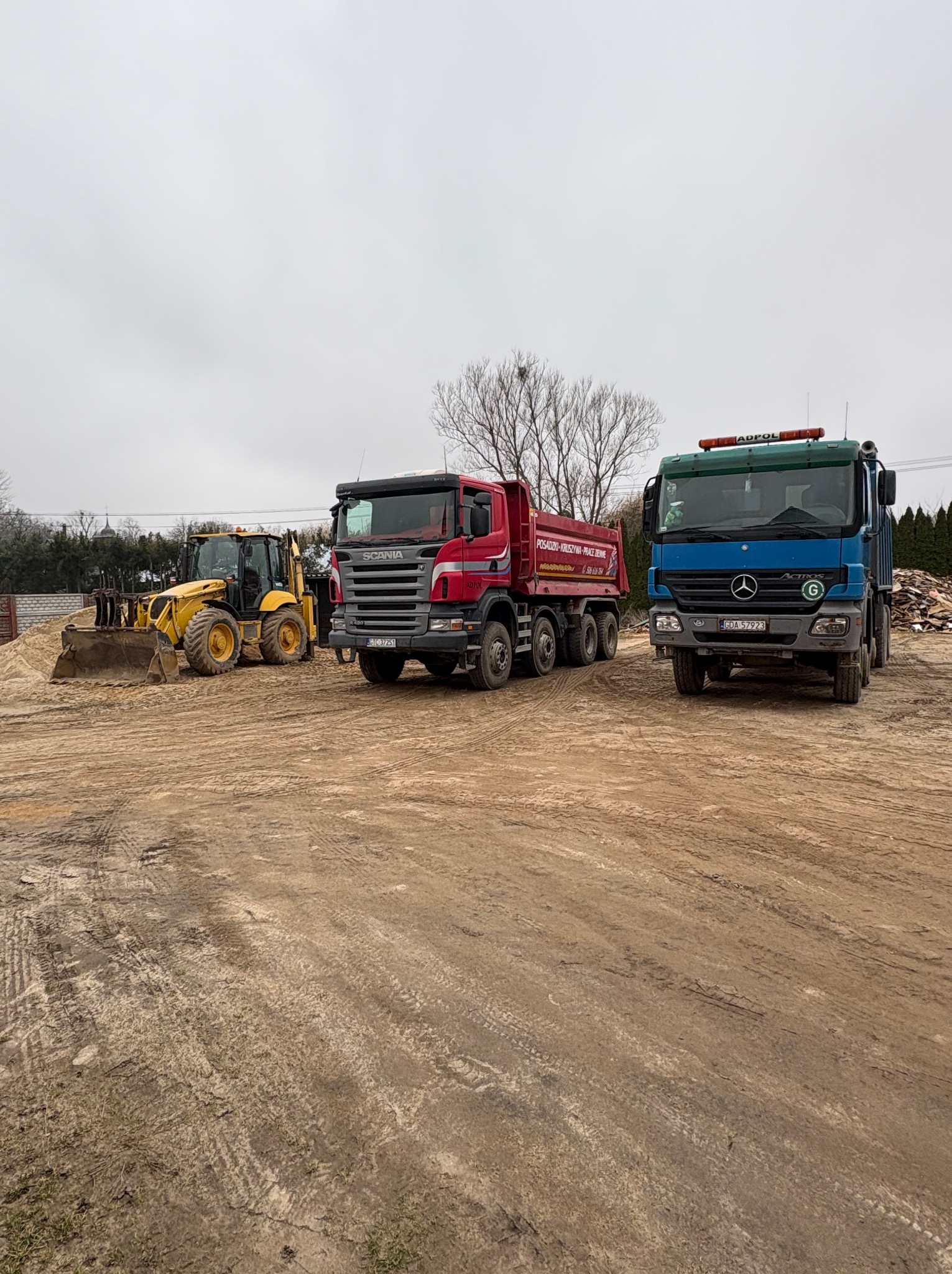 Żółta koparko-ładowarka Volvo z pełną łyżką piasku, czerwona ciężarówka Scania i niebieska ciężarówka Mercedes Actros stoją na placu budowy w pochmurny dzień.