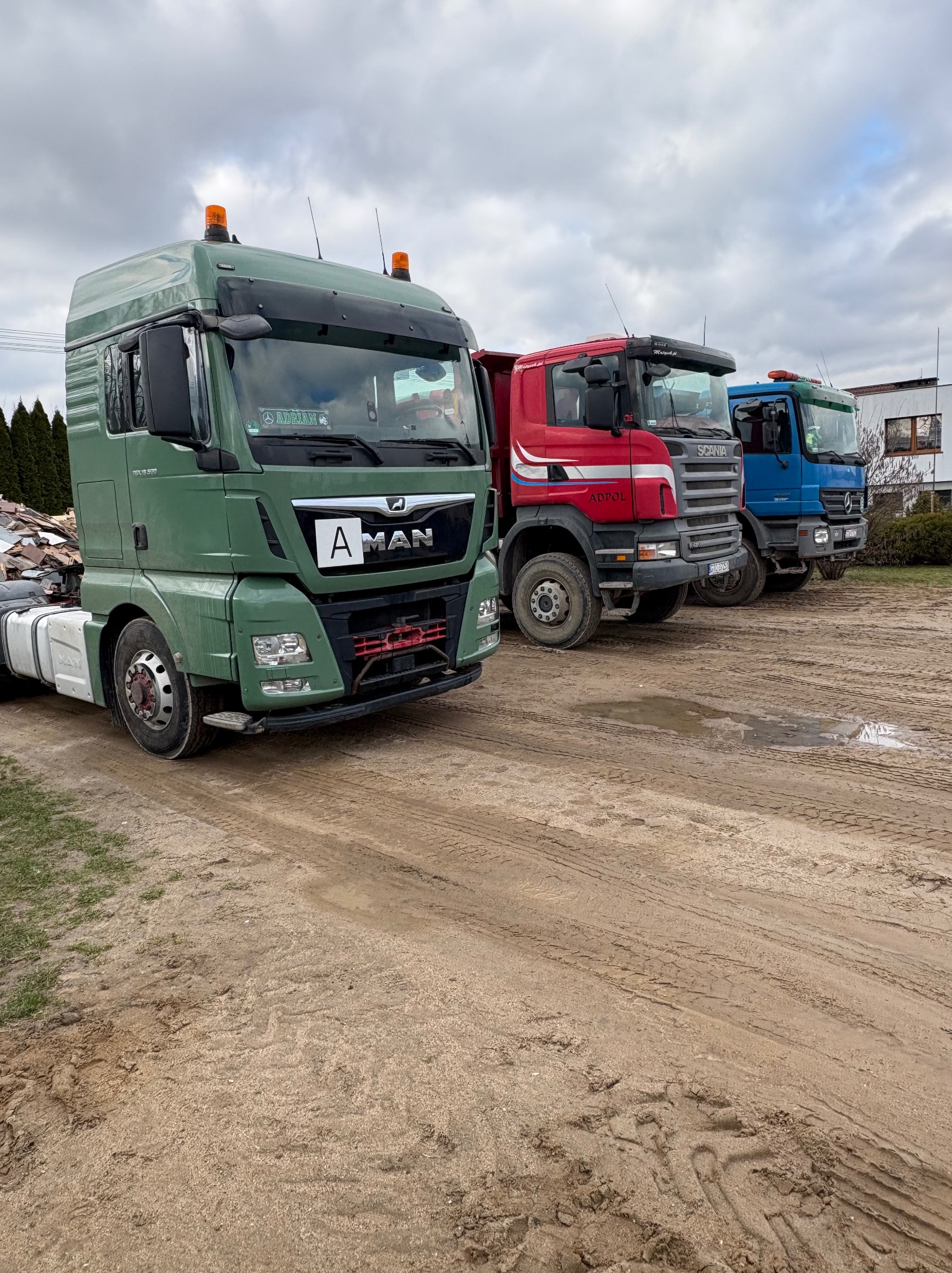 Trzy ciężarówki różnych marek i kolorów (zielona MAN, czerwona Scania, niebieska Mercedes) zaparkowane na błotnistym placu po deszczu, z widocznymi śladami opon.