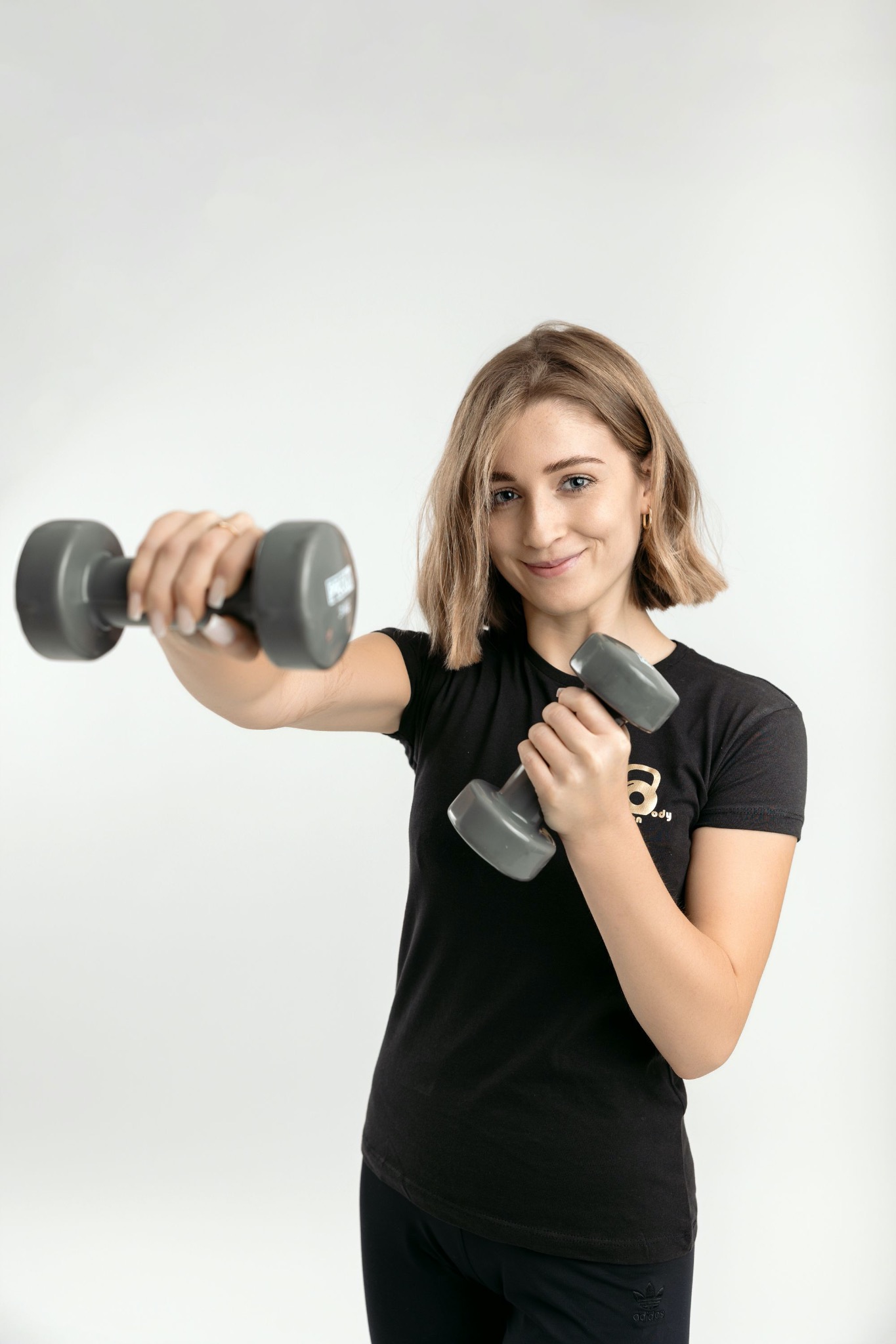 Uśmiechnięta kobieta z krótkimi blond włosami, w czarnej koszulce z logo, trzyma dwie szare hantle na białym tle.