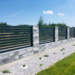 Prywatne - Nowoczesne ogrodzenie panelowe z podmurówką z szarej kostki, oddzielające trawnik od rabaty z białym żwirem i niskimi roślinami, na tle błękitnego nieba.