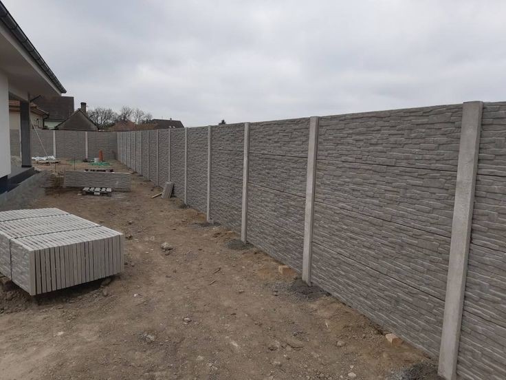 Nowoczesne, betonowe ogrodzenie panelowe o teksturze kamienia, z widocznymi słupkami i ułożonymi panelami w tle, w trakcie montażu na posesji.