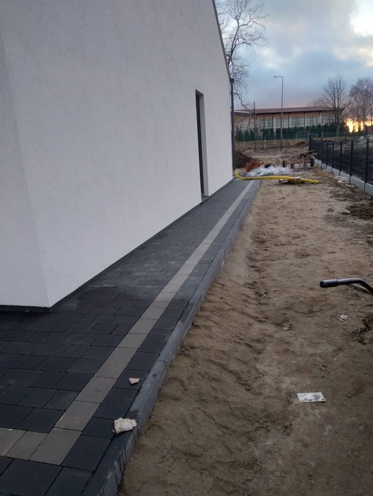 Ułożona, grafitowa kostka brukowa z szarym pasem przy białej ścianie budynku. W tle widoczne ogrodzenie i budynek. Prace brukarskie.