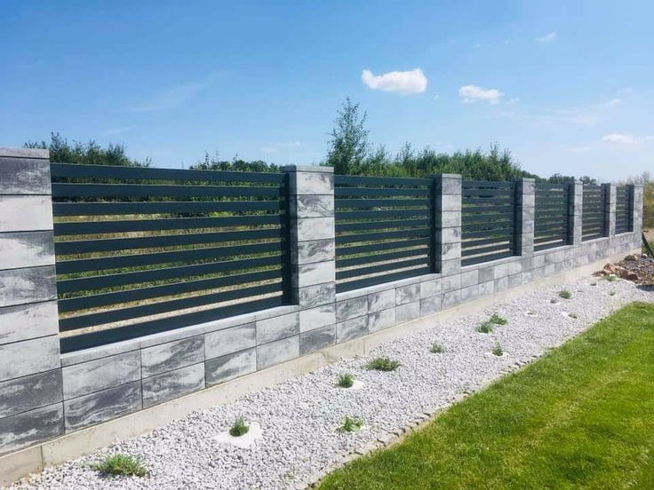Nowoczesne ogrodzenie panelowe z podmurówką z szarej kostki, oddzielające trawnik od rabaty z białym żwirem i niskimi roślinami, na tle błękitnego nieba.