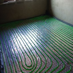 HYDRO-KLIMA Artur Kacprzak - Zielone rurki ogrzewania podłogowego ułożone na srebrnej folii z czerwoną siatką, przymocowane czarnymi klipsami, widok z góry, surowy beton na ścianie w tle.