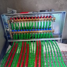 HYDRO-KLIMA Artur Kacprzak - Rozdzielacz ogrzewania podłogowego w metalowej szafce, z widocznymi numerowanymi zaworami, zielonymi rurami i czerwonymi otulinami, zamontowany na folii z rastrem.
